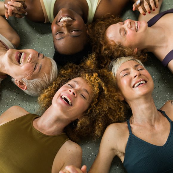 five women from different background, holding hands and laughing in pilates studio
