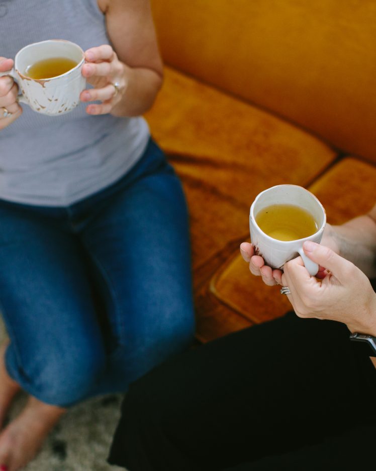 two women sit on a couch and talk together over cups of herbal tea.