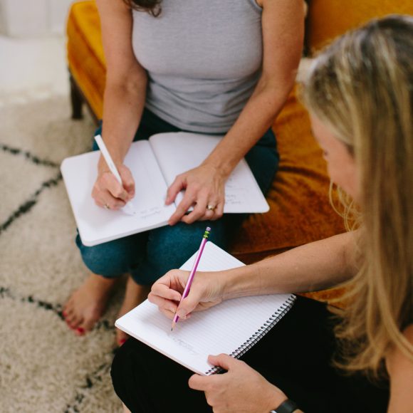 two women sit on a couch together and write in journals.
