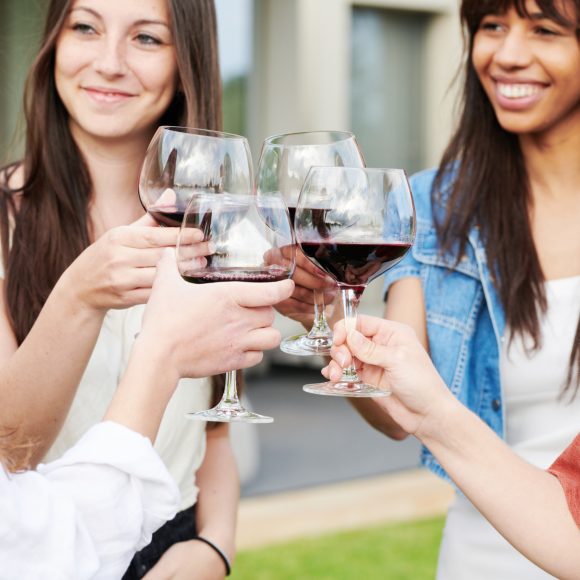 group of beautiful women celebrating together with glasses filled with red wine in summer backyard in the afternoon. They are smiling and toasting with glasses.