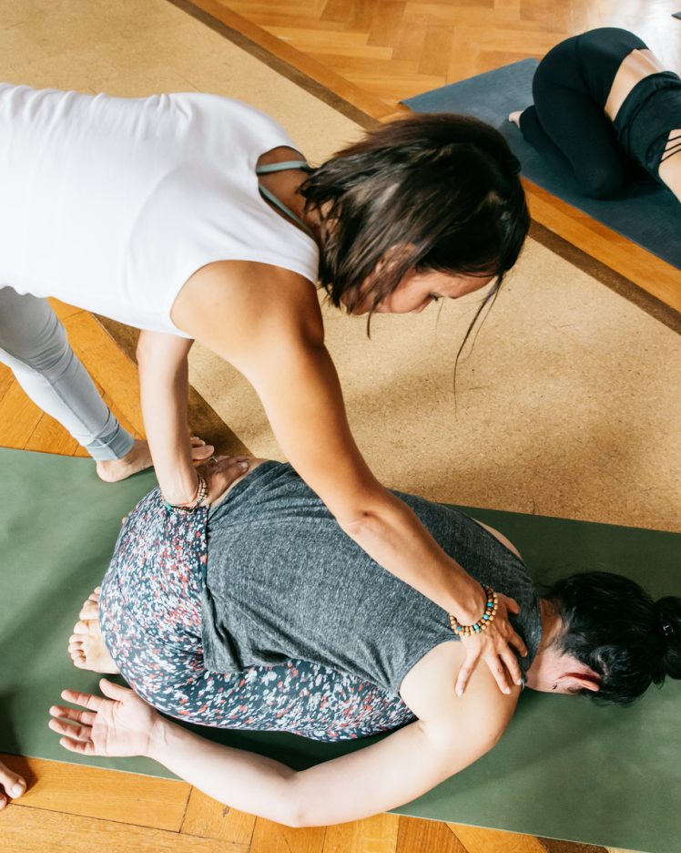 An indoor group yoga class taught by a female yoga instructor. She is helping a student with a posture. Shot in a spacious room with beautiful parquet floor and ornate ceiling.