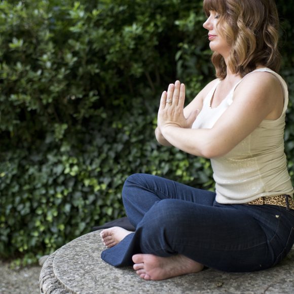 Kristal sitting outside cross legged atop a rock in a meditation pose with her hands pressed together in front of her chest.